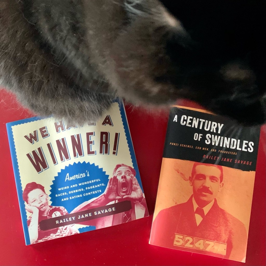 a cat casually sits on two books: We Have a Winner! and A Century of Swindles