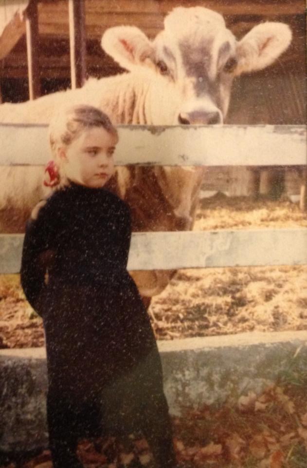 a grainy snapshot of a young girl in a dark dress looking wary of the dairy cow standing behind her