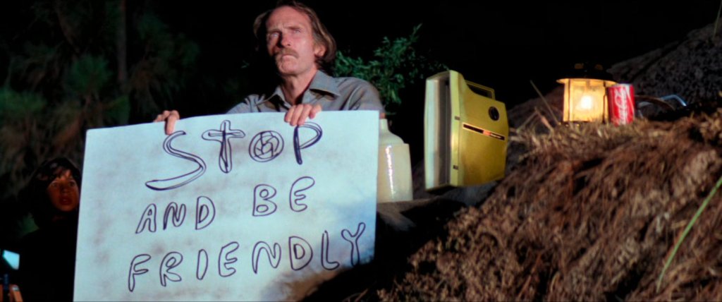 an old man sits in the dirt with a sign that reads, Stop and be friendly; from Steven Spielberg's Close Encounters of the Third Kind